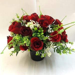 May include: A floral arrangement of deep red roses, green leaves, and small white flowers in a black pot. The artificial flowers are arranged to create a full, round display, suitable for home decor or gifting.