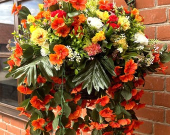 May include: A hanging basket filled with a dense arrangement of artificial flowers in vibrant shades of orange, yellow, and white, accented by green foliage and trailing vines, suspended by a metal chain against a red brick wall background.