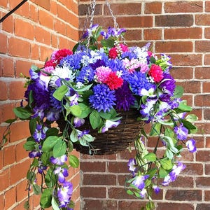 May include: A hanging basket filled with artificial flowers in shades of red, blue, purple, and white. Trailing vines of purple and white flowers cascade from the basket. The basket is made of woven brown material and hangs against a red brick wall.