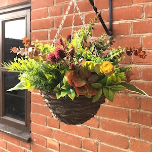 May include: A brown wicker hanging basket filled with artificial greenery, including green leaves, red and orange flowers, and yellow foliage.