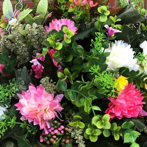 May include: A hanging basket arrangement featuring artificial flowers in shades of pink, white, and yellow, interspersed with various green foliage, small sprigs of lavender, and dark green leaves, suspended by silver metal chains against a brick wall.