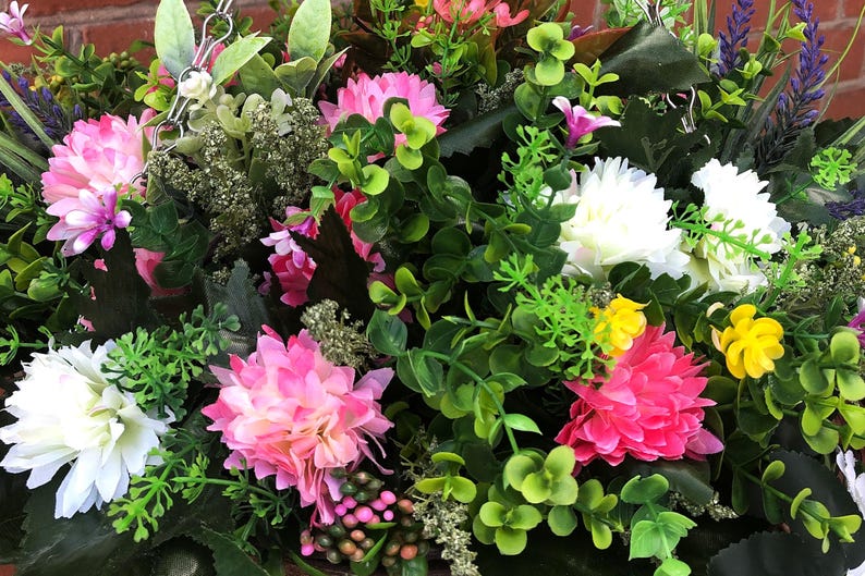 May include: A hanging basket arrangement featuring artificial flowers in shades of pink, white, and yellow, interspersed with various green foliage, small sprigs of lavender, and dark green leaves, suspended by silver metal chains against a brick wall.