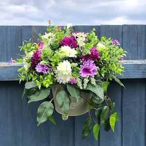 May include: A hanging floral display with purple, white, and fuchsia flowers and green foliage. The flowers are arranged in a light green metal pot, suspended on a blue wooden fence. The arrangement adds a touch of color.