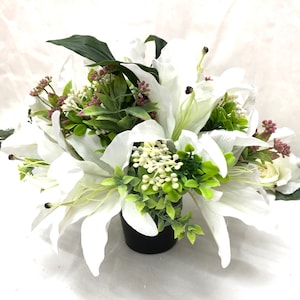 May include: A white artificial lily arrangement in a black pot. The lilies are surrounded by green foliage and small white flowers.