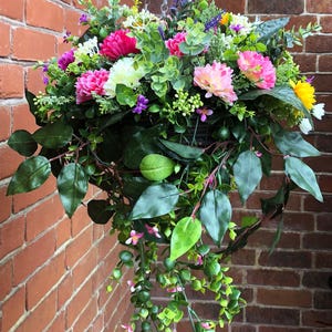 May include: A hanging basket filled with an arrangement of artificial flowers including pink, white, and yellow blossoms, interspersed with green foliage and trailing vines. The display is suspended by a silver-toned metal chain.