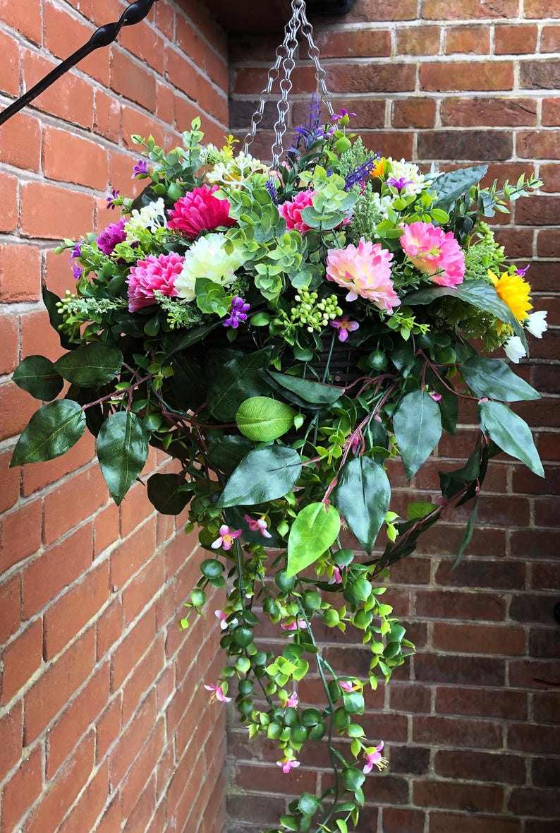 May include: A hanging basket filled with an arrangement of artificial flowers including pink, white, and yellow blossoms, interspersed with green foliage and trailing vines. The display is suspended by a silver-toned metal chain.