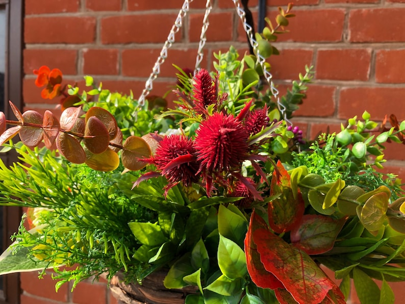May include: A hanging basket filled with a variety of artificial plants and flowers. The basket is made of woven material and features a mix of green foliage, red flowers, and brown leaves.