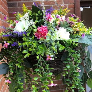 May include: A hanging basket filled with a variety of artificial flowers and greenery. The arrangement includes white, pink, and yellow blossoms mixed with purple sprigs and trailing green vines, all contained within a dark brown woven wicker basket.