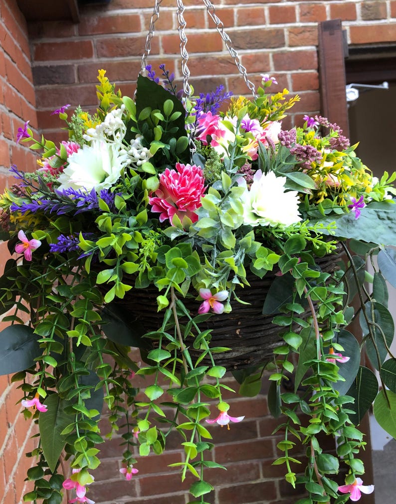 May include: A hanging basket filled with a variety of artificial flowers and greenery. The arrangement includes white, pink, and yellow blossoms mixed with purple sprigs and trailing green vines, all contained within a dark brown woven wicker basket.