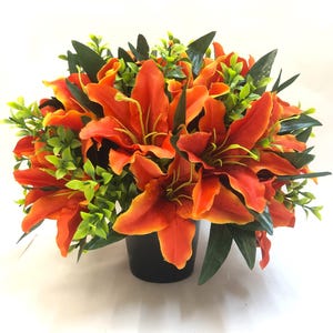 May include: A colourful floral display of orange lilies and green foliage. The lilies, with their large petals and dark centres, are the main feature. The arrangement is presented in a black pot, providing a strong contrast to the bright colours.