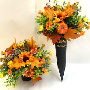 May include: Two artificial floral arrangements featuring orange sunflowers, maple leaves, green foliage, and small pumpkins. One arrangement is in a black cone-shaped stake with the gold text In Loving Memory, and the other is in a black pot.