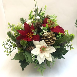May include: A festive Christmas floral arrangement featuring red and white poinsettias, green foliage, and gold-painted pinecones. The arrangement includes sprigs of greenery and small white berries, creating a holiday centerpiece.