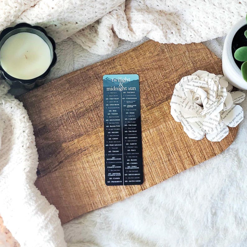 May include: A bookmark with the text "Twilight & Midnight Sun" and a list of chapter titles. The bookmark is on a wooden cutting board with a candle and a paper flower. The background includes a white knitted blanket and a small potted plant.