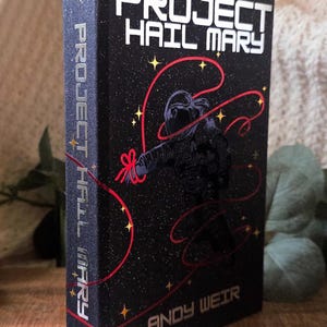 May include: Hardcover book titled "Project Hail Mary" by Andy Weir. The cover is black with white text and a stylized astronaut illustration with red lines and stars. The spine also features the title.