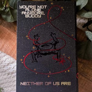 May include: Black book cover with the text "YOU'RE NOT ALONE ANYMORE, BUDDY" and "NEITHER OF US ARE" in gold lettering. A black illustration of a frog-like creature is featured, with red string and gold star accents.
