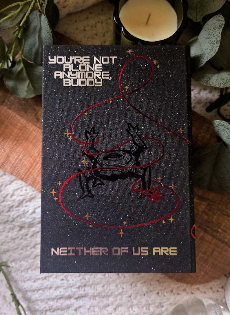 May include: Black book cover with the text "YOU'RE NOT ALONE ANYMORE, BUDDY" and "NEITHER OF US ARE" in gold lettering. A black illustration of a frog-like creature is featured, with red string and gold star accents.
