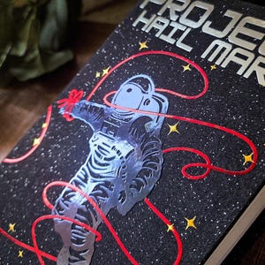 May include: A hardcover book titled "PROJECT HAIL MARY" with a black cover featuring a silver astronaut design and red lines. The background is a starry night with yellow stars. The book's title is in silver lettering.