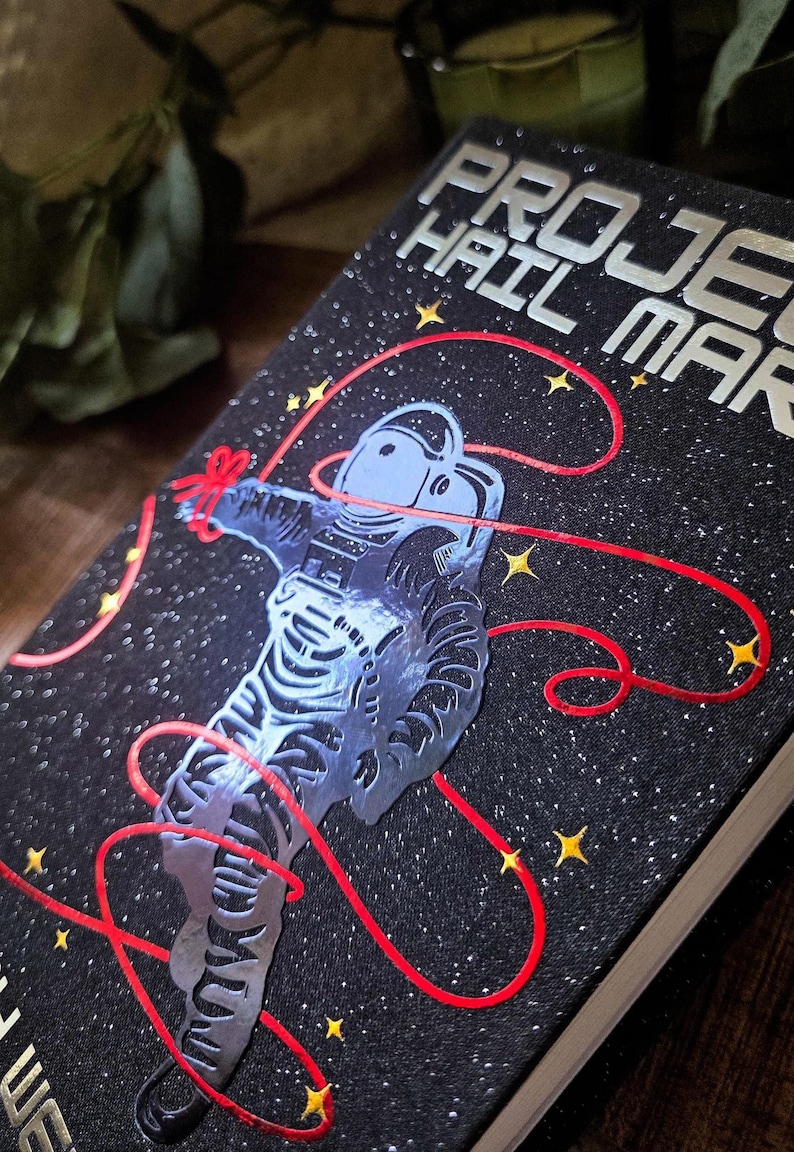 May include: A hardcover book titled "PROJECT HAIL MARY" with a black cover featuring a silver astronaut design and red lines. The background is a starry night with yellow stars. The book's title is in silver lettering.