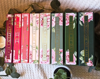 May include: A collection of hardcover books with floral designs and gold lettering. Titles include "The Assassin's Blade," "Throne of Glass," and "Kingdom of Ash." The spines feature pink roses and green leaves, creating a romantic aesthetic.
