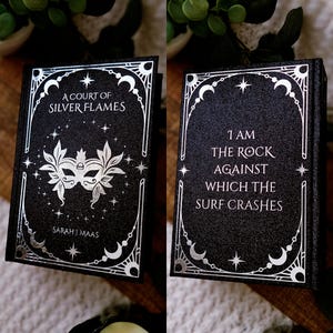 May include: A black book with silver foil detailing. The front cover reads "A Court of Silver Flames" and features a mask design. The back cover has the quote, "I am the rock against which the surf crashes."