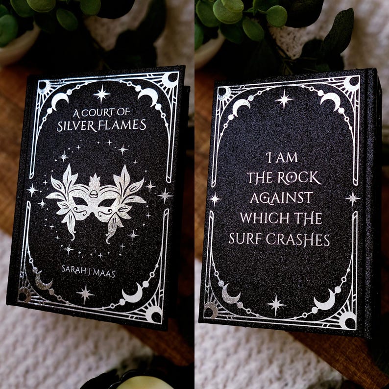 May include: A black book with silver foil detailing. The front cover reads "A Court of Silver Flames" and features a mask design. The back cover has the quote, "I am the rock against which the surf crashes."