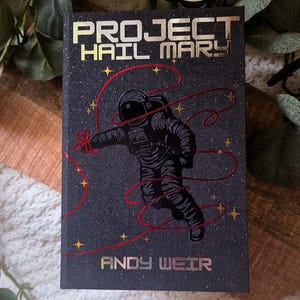 May include: A hardcover book titled "PROJECT HAIL MARY" by Andy Weir. The cover features a black background with a textured, glittery effect, an astronaut illustration, and gold lettering. Red lines and gold stars accent the design.