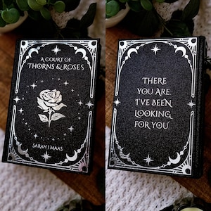 May include: A black book with silver foil detailing. The front cover reads "A Court of Thorns & Roses" with a rose illustration. The back cover says "There you are. I've been looking for you."