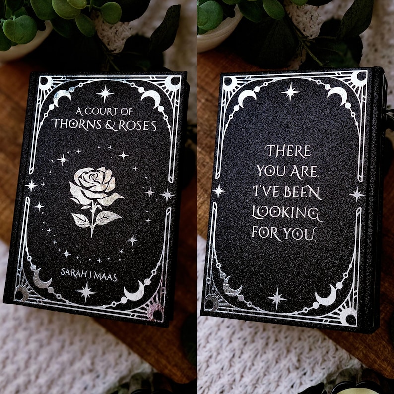 May include: A black book with silver foil detailing. The front cover reads "A Court of Thorns & Roses" with a rose illustration. The back cover says "There you are. I've been looking for you."