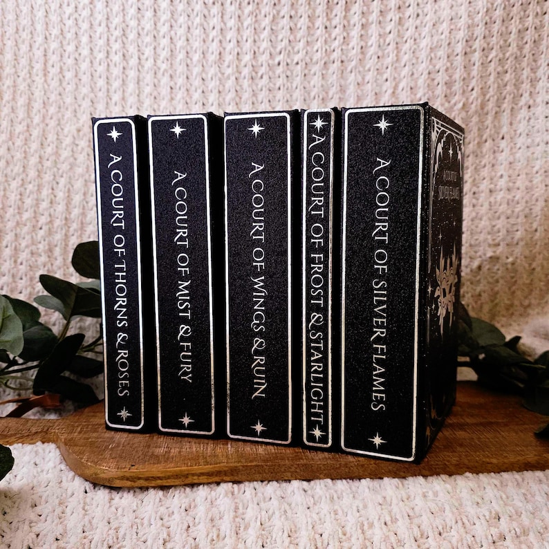 May include: A set of black books with white lettering, featuring titles such as "A Court of Thorns & Roses" and "A Court of Silver Flames." The books are displayed on a wooden board, set against a neutral backdrop.