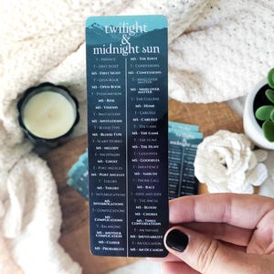 May include: A bookmark with the text "twilight & midnight sun" and a list of chapter titles. The bookmark is held in a hand with a black painted fingernail. The background is a cream-colored, textured surface.
