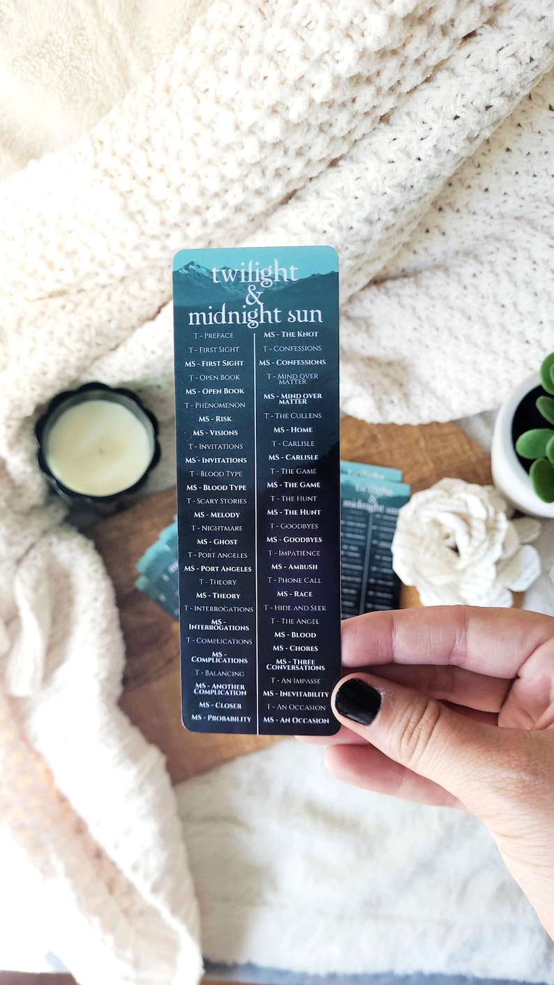 May include: A bookmark with the text "twilight & midnight sun" and a list of chapter titles. The bookmark is held in a hand with a black painted fingernail. The background is a cream-colored, textured surface.