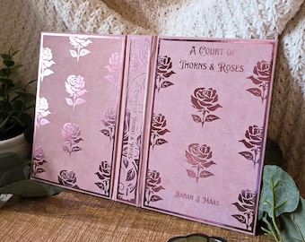 May include: A pink book with the title "A Court of Thorns & Roses" and the author's name, Sarah J. Maas, is displayed. The cover features rose illustrations in a darker pink. The book is open, revealing the spine and inner pages.