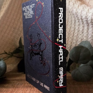 May include: A hardcover book with a dark blue cover featuring the title "Project Hail Mary" in white lettering on the spine. The front cover has the text "You're Not Alone Anymore, Buddy" and a red line design. The book is on a wooden surface.