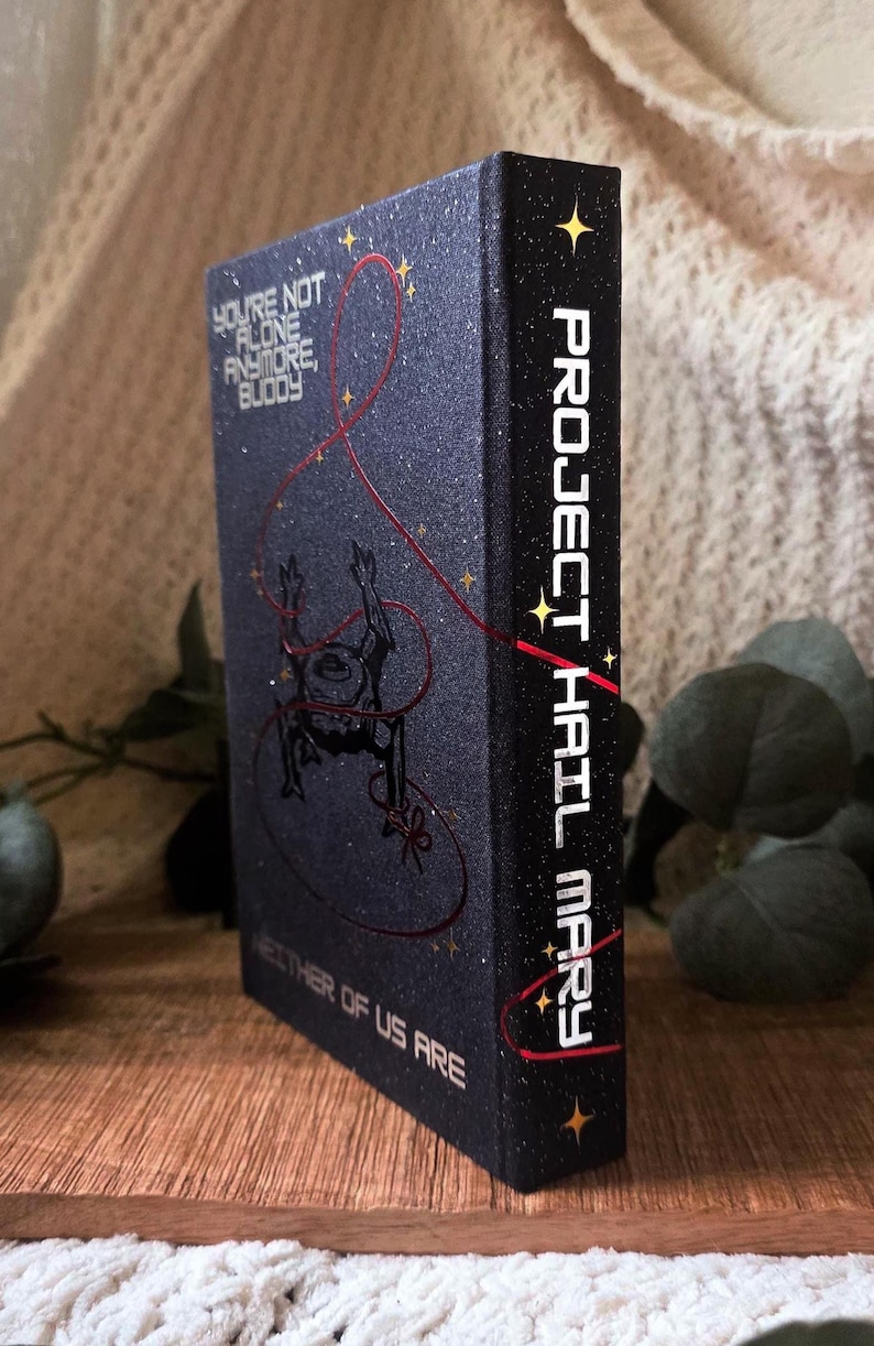 May include: A hardcover book with a dark blue cover featuring the title "Project Hail Mary" in white lettering on the spine. The front cover has the text "You're Not Alone Anymore, Buddy" and a red line design. The book is on a wooden surface.