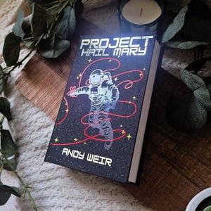 May include: A hardcover book titled "PROJECT HAIL MARY" by Andy Weir. The cover features an astronaut illustration with red lines against a starry black background. The book is resting on a wooden surface.