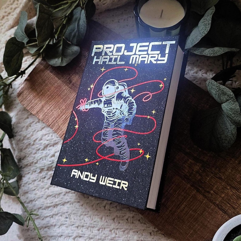 May include: A hardcover book titled "PROJECT HAIL MARY" by Andy Weir. The cover features an astronaut illustration with red lines against a starry black background. The book is resting on a wooden surface.