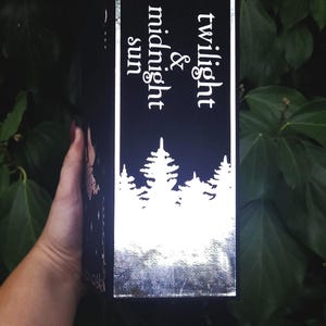 Może przedstawiać: Czarne pudełko z srebrną obw&oacute;dką i tekstem "twilight & midnight sun" w białych literach. Pudełko przedstawia białą sylwetkę lasu z drzewami.