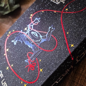 May include: A black book with a starry design and the text "YOU'RE NOT ALONE ANYMORE, BUDDY." A blue alien-like figure is connected by a red line to a red bow. Yellow stars are scattered across the cover.