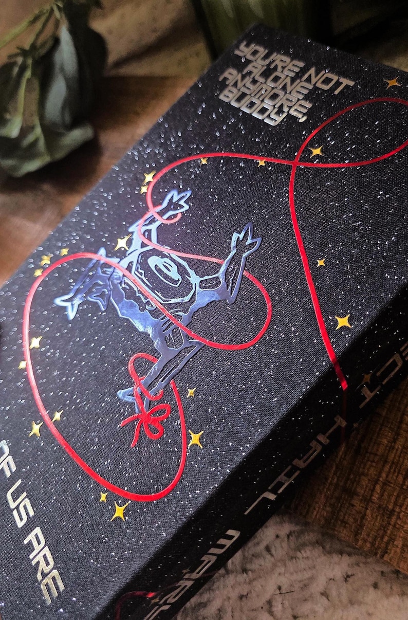 May include: A black book with a starry design and the text "YOU'RE NOT ALONE ANYMORE, BUDDY." A blue alien-like figure is connected by a red line to a red bow. Yellow stars are scattered across the cover.