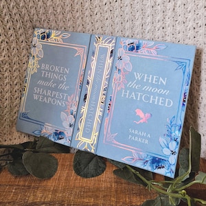 May include: A light blue book with the title "When the Moon Hatched" and the author's name, Sarah A. Parker. The cover features floral designs and the text "Broken Things Make the Sharpest Weapons". The book is open, revealing the spine.