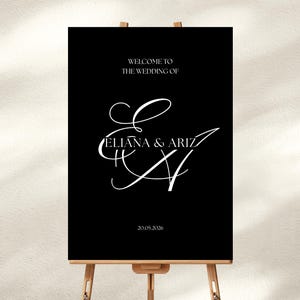 Könnte beinhalten: Ein schwarzes Hochzeits-Willkommensschild mit weißer, eleganter Schrift. Auf dem Schild steht "WELCOME TO THE WEDDING OF ELIANA & ARIZ" und das Datum "20.05.2026". Das Schild wird auf einer Holzstaffelei präsentiert.