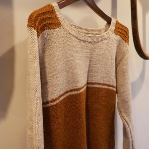 May include: A beige and brown knitted jumper with a round neckline and long sleeves. The jumper has a brown stripe across the bottom and brown accents on the shoulders.