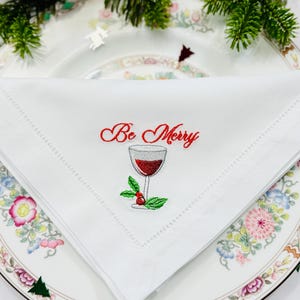 Könnte beinhalten: Weißes Leinen-Servietten mit der roten Stickerei "Be Merry" und einer Weinglas-Illustration. Die Serviette liegt auf einem dekorativen Teller mit floralen und grünen Bordüren.