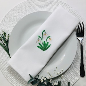 May include: White linen napkin with an embroidered design of white snowdrops with green leaves on a white plate with a white woven placemat.