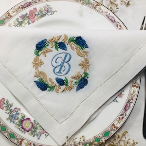 May include: A white linen napkin with a blue and gold embroidered wreath and the letter "B" in the centre.