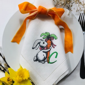 May include: White linen napkin embroidered with a bunny clutching a carrot, tied with a pink bow. The napkin is folded and decorated with an orange velvet ribbon. A white plate, black fork, and yellow daffodils are in the background.