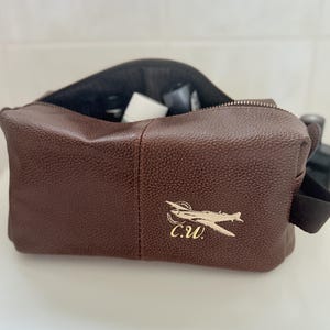 May include: A brown leather wash bag with a zip closure. The bag has a gold embroidered airplane design and the initials "C.W." on the front. The bag is open, showing the interior and some toiletries.