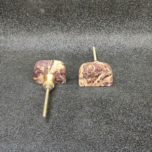 May include: Two decorative cabinet knobs with a marbled brown and beige pattern. Each knob has a brass stem and a square-shaped face. One knob has a screw attached, and the other has the word "craftmuse" written on it.