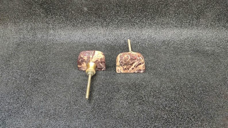 May include: Two decorative cabinet knobs with a marbled brown and beige pattern. Each knob has a brass stem and a square-shaped face. One knob has a screw attached, and the other has the word "craftmuse" written on it.