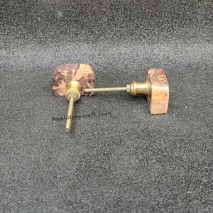 May include: Two decorative cabinet knobs. Each knob features a rectangular, brown and tan stone face with a brass stem and hardware. The knobs are designed for furniture and home decor.